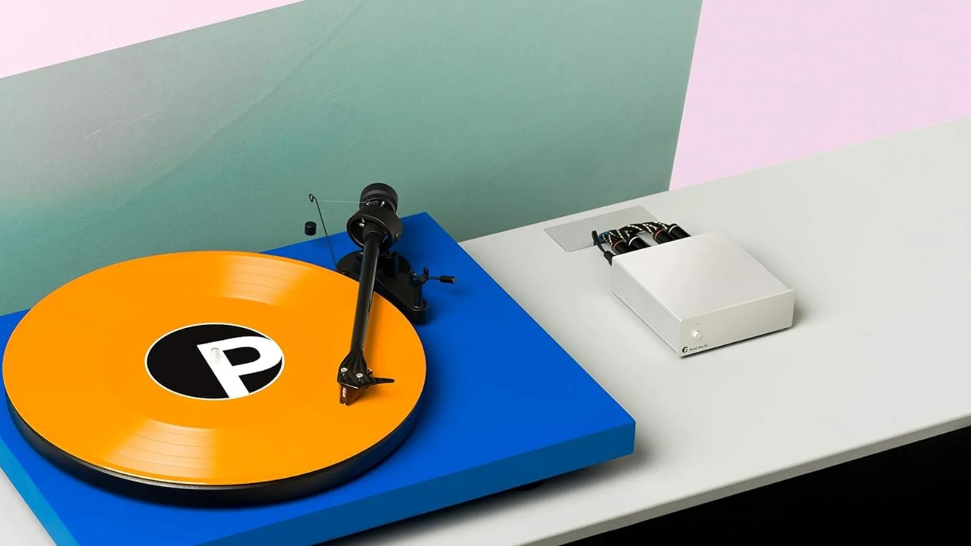 Pro-Ject Phono Box S2 en situation d'écoute, branché à une platine Pro-Ject Primary E bleue avec disque vinyle orange sur fond rose et vert pastel